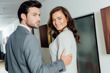 confident businessman touching hand of cheerful businesswoman in formal wear while standing in hotel corridorの写真素材