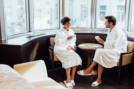 cheerful woman and handsome man in bathrobes looking at each other while holding cups of coffeeの写真素材