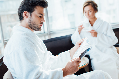 selective focus of handsome man reading newspaper in bathrobe near girlfriend holding cupの写真素材