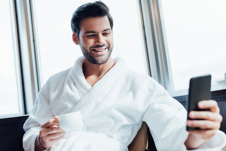 selective focus of cheerful bearded man in white bathrobe using smartphone and holding cup with coffeeの写真素材