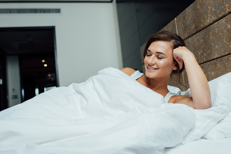 cheerful brunette woman smiling while lying on pillows with closed eyesの写真素材