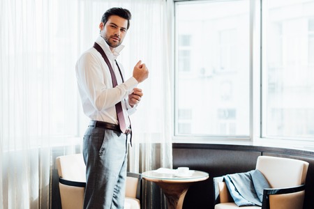 confident bearded man in suit touching shirt while standing in hotel