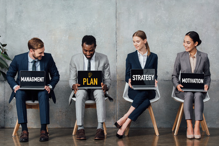 smiling multiethnic businesspeople sitting on chairs and holding laptops with plan, strategy,...