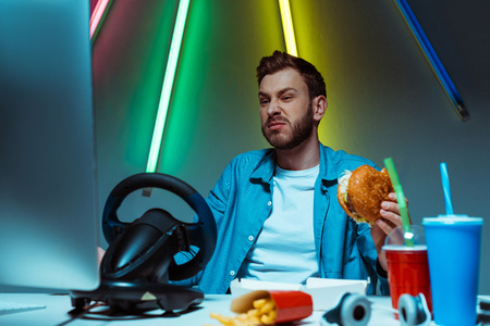 selective focus of handsome man eating burger and looking at computer monitor