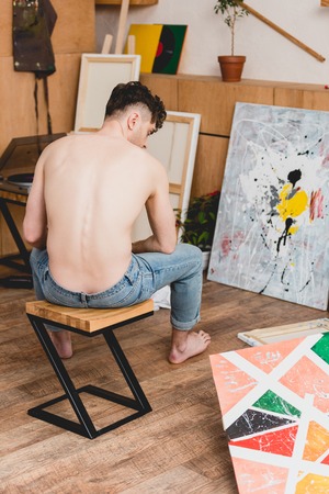 half-naked artist in blue jeans sitting on chair in gallery, surrounded by paintingsの写真素材
