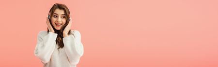 panoramic shot of smiling and beautiful woman in white sweater listening music with headphones isolated on pinkの写真素材