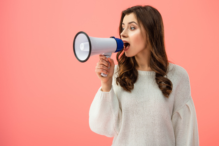 brunette woman in white sweater screaming in megaphone isolated on pink