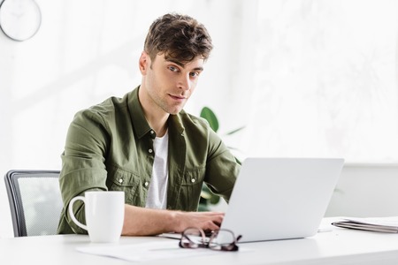 man in green shirt sitting at table with laptop and typing in office