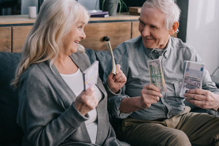 happy senior couple sitting on couch and holding euro and dollar banknotesの写真素材