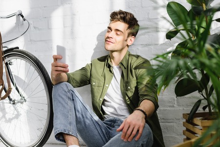 handsome man in green shirt holding smartphone, sitting near brick wall, bicycle and plants in officeの写真素材