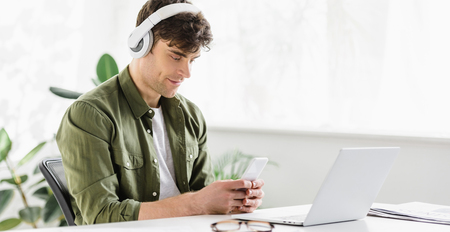 handsome businessman in headphones sitting near table with laptop and holding smartphone in officeの写真素材