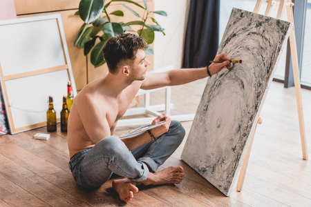 handsome half-naked artist sitting with crossed legs on floor and painting picture on canvasの写真素材
