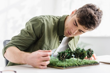 handsome architect sitting at table and putting solar panels models on grass in officeの写真素材