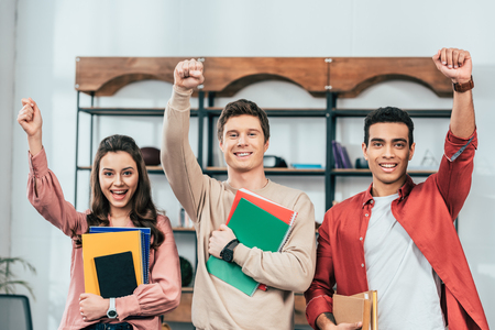 Three cheerful multiethnic students holding notebooks and books and holding fists upの写真素材