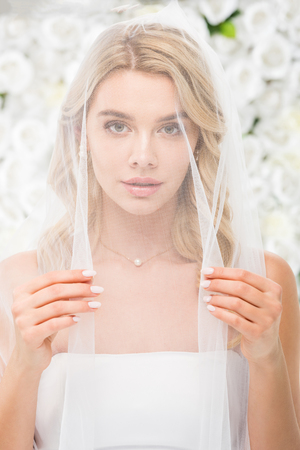 beautiful young woman with face covered with transparent bridal veil looking at camera on white floral backgroundの写真素材