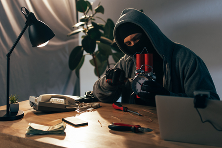 Criminal in mask sitting at table with laptop and making bombの写真素材