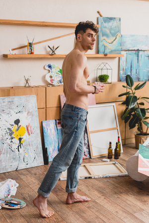 handsome half-naked artist in blue jeans standing in painting studio and looking awayの写真素材