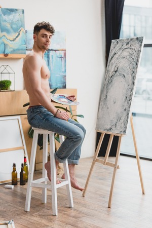 good-looking half-naked artist looking at camera while sitting on high chair near easel with paintingの写真素材