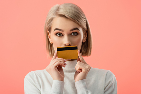 surprised blonde woman covering mouth with credit card isolated on pinkの写真素材