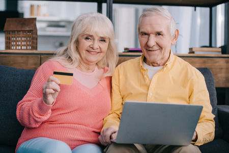 happy senior couple sitting on couch with laptop and credit card, looking at camera and doing online shopping at homeの写真素材