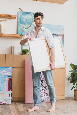 good-looking artist in white shirt and blue jeans carrying canvas in painting studioの写真素材