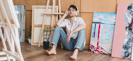 panoramic shot of handsome artist in headphones, listening music while sitting on floorの写真素材