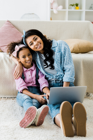 smiling african american mother and daughter using laptop while sitting on carpet near sofaの写真素材
