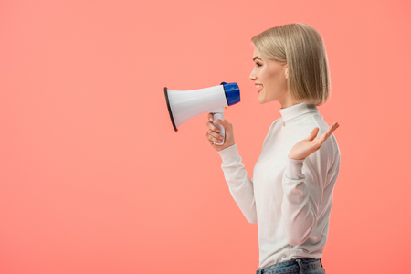 cheerful blonde woman speaking in megaphone isolated on pinkの写真素材