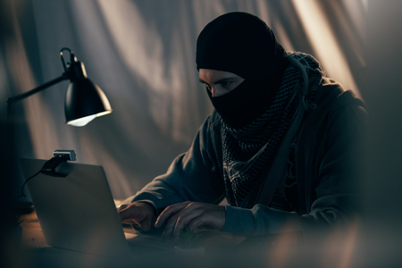 Terrorist in black mask typing on laptop keyboard in roomの写真素材