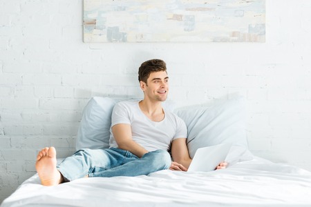 handsome man in white t-shirt and jeans sitting on bed with laptop and smiling in bedroomの写真素材