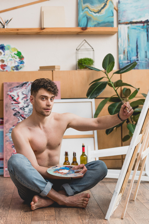 handsome half-naked artist holding palette and painting picture on canvasの写真素材