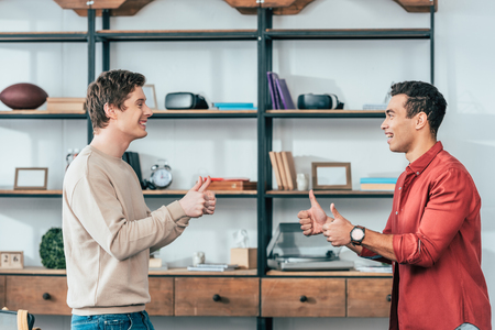 Side view of two multiethnic smiling friends showing thumbs up and looking at each otherの写真素材