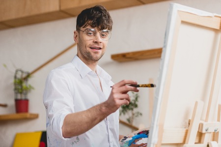 selective focus of smiling artist in white shirt and glasses painting on canvas and looking at cameraの写真素材