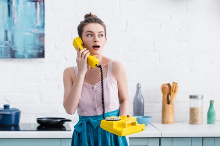 shocked young woman talking on retro yellow telephone in kitchenの写真素材