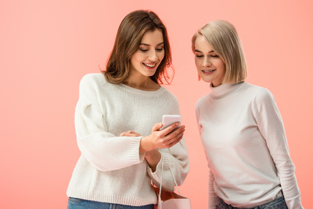 cheerful blonde and brunette friends looking at smartphone isolated on pinkの写真素材