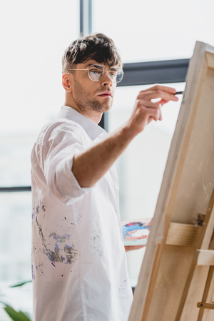 selective focus of handsome artist in glasses painting on canvasの写真素材