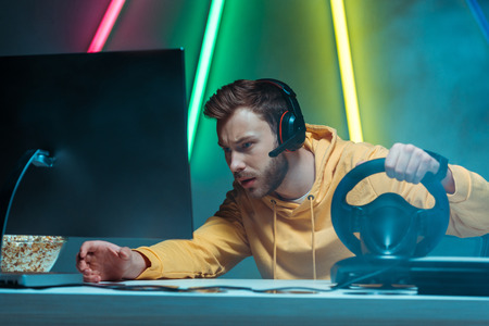 young adult and handsome man in headphones playing video game with steering wheel