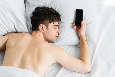 top view of handsome man sleeping under blanket in bad near smartphone with blank screen at homeの写真素材