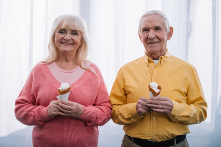 smiling senior couple looking at camera and holding ice cream cones at homeの写真素材