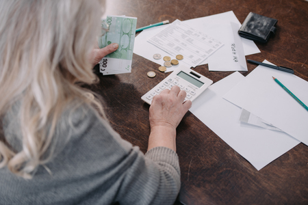 back view of senior woman sitting at table, using calculator and counting moneyの写真素材