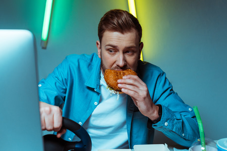handsome man playing video game with steering wheel and eating burger