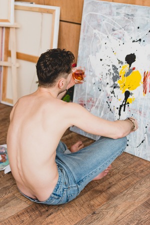 half-naked artist sitting on floor in front of picture and holding glass of whiskeyの写真素材