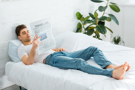 man in white t-shirt and jeans lying on bed and reading newspaper at homeの写真素材