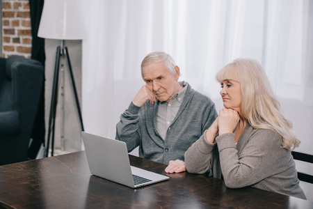 senior couple sitting at table, propping chins with hands and using laptop at homeの写真素材