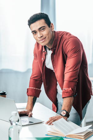 Smiling student in red shirt standing near table with laptop and looking at cameraの写真素材