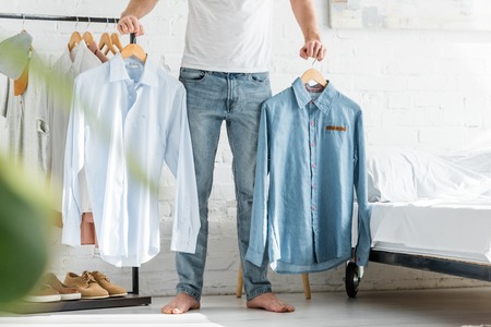 cropped view of man in white t-shirt standing near bed and holding shirts in bedroomの写真素材