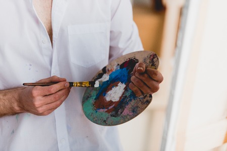 partial view of artist in white shirt mixing paints with paintbrush on palette
