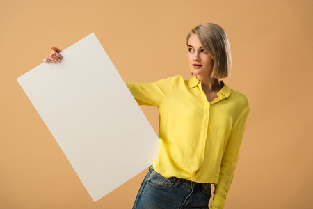 Dreamy young woman in yellow shirt holding blank placard isolated on beige