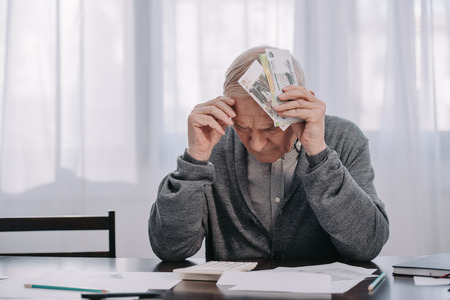stressed senior man sitting at table with paperwork and holding money at homeの写真素材