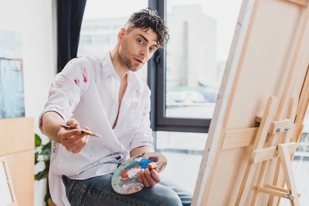 good-looking artist with palette and paintbrush looking at cameraの写真素材